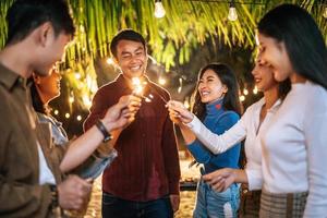 porträtt av Lycklig asiatisk grupp av vänner har roligt med sparklers utomhus- - ung människor har roligt med fyrverkeri på natt tid - människor, mat, dryck livsstil, ny år firande begrepp. foto