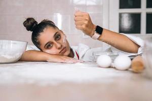 trött ung kvinna är häller mjöl på de kök tabell foto