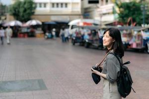 ung asiatisk kvinna ryggsäck resande använder sig av digital kompakt kamera, njuter gata kulturell lokal- plats och leende. resande kontroll ut sida gator. foto