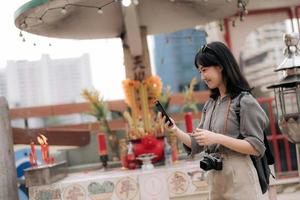 ung asiatisk kvinna ryggsäck resande använder sig av mobil telefon, njuter gata kulturell lokal- plats. resande kontroll ut sida gator. foto
