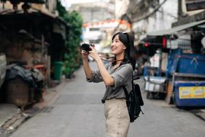 ung asiatisk kvinna ryggsäck resande använder sig av digital kompakt kamera, njuter gata kulturell lokal- plats och leende. resande kontroll ut sida gator. foto