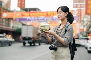 Lycklig ung asiatisk kvinna ryggsäck resande dricka en kokos juice på Kina stad gata mat marknadsföra i Bangkok, thailand. resande kontroll ut sida gator. foto