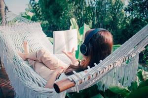 ung avslappnad kvinna lyssnande till musik med hörlurar läsning bok på vit hängmatta i trädgård på Hem. morgon- Sol. långsam levande, helgen fritid aktivitet. karantän och själv isolering period. foto