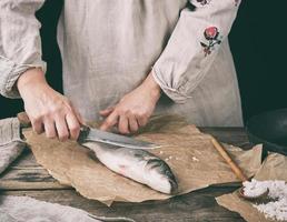 kvinna i grå Linné kläder rengör de fisk hav bas skalor på en brun trä- styrelse foto