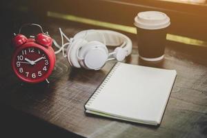 kopp kaffe, anteckningsbok och röd väckarklocka med hörlurar på ett träbord