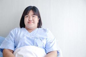 fett asiatisk kvinna patient med ljus leende sitta i en sjukhus säng. begrepp av hälsa problem för fet människor, hälsa försäkring. kopia Plats foto