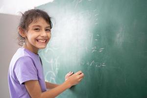 Lycklig skol porträtt av liten flicka skrivning på grön svarta tavlan i klassrum, utbildning och skola begrepp foto