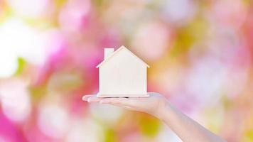 kvinna hand innehav små Hem modell trä- med rosa bokeh abstrakt bakgrund. familj liv och företag verklig egendom koncept, eko hus foto