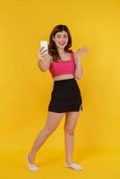 porträtt av leende ung kvinna selfie med mobil telefon i henne händer medan stående isolerat över gul bakgrund foto