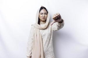 besviken asiatisk muslim kvinna bär en hijab ger tummen ner hand gest av ogillande, isolerat förbi vit bakgrund foto