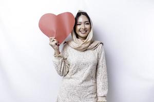 en Lycklig ung asiatisk muslim kvinna bär en hijab känner romantisk former hjärta gest uttrycker anbud känslor och innehav en röd hjärtformade papper foto