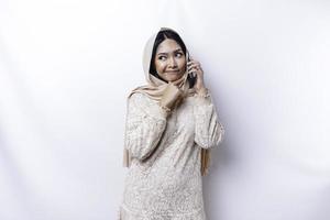 en omtänksam ung asiatisk muslim kvinna bär en hijab och innehav henne haka medan talande på de telefon isolerat förbi en vit bakgrund foto