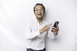 upphetsad asiatisk man bär vit skjorta leende medan innehav hans telefon, isolerat förbi vit bakgrund foto