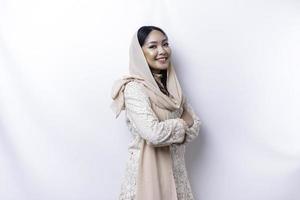 porträtt av en självsäker leende asiatisk muslim kvinna bär hijab stående med vapen vikta och ser på de kamera isolerat över vit bakgrund foto