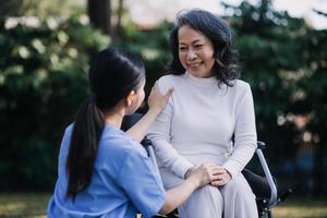 asiatisk ung vårdgivare sjuksköterska Stöd senior äldre manlig gående utomhus. specialist flicka läkare hjälp och ta vård av äldre mogna man patient håller på med fysisk terapi i offentlig parkera på amning Hem. foto