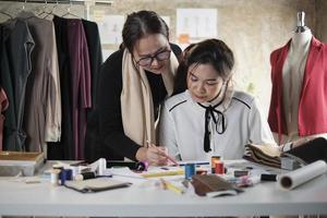 asiatisk medelålders kvinna mode designer lär en ung tonåring praktikant skräddare i studio med färgrik tråd och sömnad tyg för klänning design samling idéer, professionell boutique små företag. foto