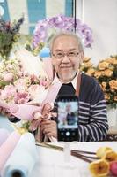 e-handel företag, ett senior manlig blomsterhandlare arbetare demonstrera och visa blommig arrangemang via uppkopplad leva strömning med smartphone Ansökan i en ljus blomma affär, en skön blommar Lagra. foto