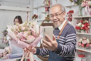 Lycklig asiatisk senior manlig blomsterhandlare ägare selfie bilder och visa blommig bukett arrangemang med smartphone i valentine på ljus blomma affär, skön blommar Lagra, små företag sme entreprenör. foto