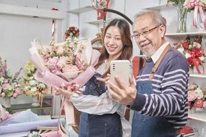 e-handel företag, senior asiatisk manlig blomsterhandlare arbetstagare och dotter visa blommig arrangemang, uppkopplad leva strömning, selfie förbi smartphone Ansökan i ljus blomma affär, skön blommar Lagra. foto