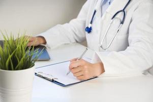 oigenkännlig kvinna läkare i vit medicinsk enhetlig sitta på skrivbord håller på med pappersarbete. sjukvård begrepp foto