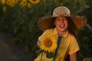 en Lycklig ung flicka med lång hår i en sugrör hatt står i en stor fält av solrosor. sommar dag. en värma solnedgång foto