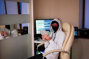 pojke hacker med pengar dollar. internet stöld . man bär en Balaklava sat Bakom en bärbar dator. foto