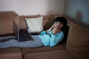 ung tonåring pojke i främre av en bärbar dator på en säng på kväll. foto