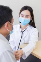 asiatisk man patient är kontrollerade upp hans hälsa medan en kvinna läkare använda sig av en stetoskop till höra hjärta Betygsätta av honom i coronavirus pandemi förbi bär en kirurgisk mask på Allt gånger. coronavirus skydd. foto