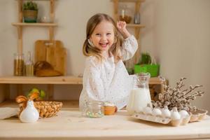 söt liten flicka i en bomull klänning på Hem i en trä- kök förbereder ett påsk kaka foto