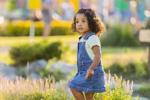 porträtt liten Lycklig lekfull litet barn svartmuskig flicka i en denim sundress stående i de trädgård på en solig dag. gående i de färsk luft. begrepp av en Lycklig barndom. Plats för text. hög kvalitet Foto