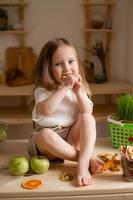 söt liten flicka äter naturlig pastill på Hem i en trä- kök. mat för barn från naturlig Produkter foto