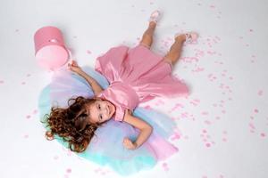 skön liten flicka i en chic rosa klänning poser Sammanträde på en grå bakgrund och kastar konfetti. mode och stil. Plats för text. Foto i de studio. hög kvalitet Foto