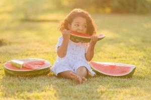 en liten svartmuskig flicka med lockigt hår i en blek rosa klänning äter en vattenmelon på de gräsmatta. picknick i de parkera. Lycklig barndom. Plats för text. hög kvalitet Foto