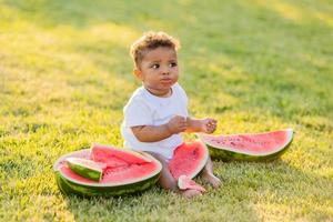 en liten svartmuskig flicka i vit kläder äter en vattenmelon på en grön gräsmatta. picknick i de parkera. Lycklig barndom. Plats för text. hög kvalitet Foto