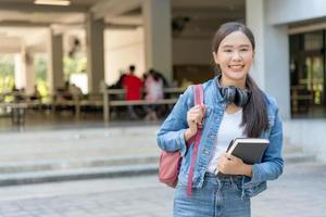 skön studerande asiatisk kvinna med ryggsäck och böcker utomhus. leende flicka Lycklig bärande en massa av bok i högskola campus. porträtt kvinna på internationell Asien universitet. utbildning, studie, skola foto