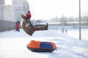 en Lycklig pojke upp i de luft på en rör sledding i de snö.. en pojke diabilder ner en kulle i vinter. foto