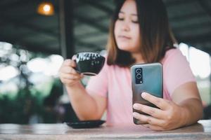 Lycklig asiatisk kvinna innehav och använder sig av mobil telefon i kaffe affär, njuter de smak av kaffe. eller kaffe affär med kopia Plats foto