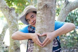 stilig asiatisk man botaniker kramar trunk av träd i skog, göra tecken av hjärta form. begrepp, kärlek natur och skydda, skog ,ekologi och miljö bevarande. . foto