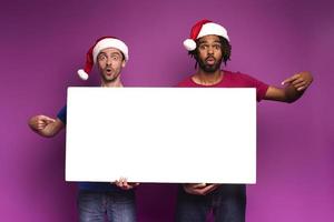 överraskad förvånad vänner med jul hatt håll en vit baner för din meddelande. lila bakgrund foto