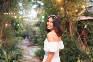 skön ung asiatisk kvinna i vit klänning sväng av ansikte leende i lysande tropisk trädgård foto