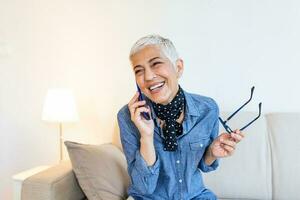 Lycklig senior kvinna använder sig av mobil telefon på Hem. leende Häftigt gammal kvinna med vit hår Sammanträde på soffa och meddelandehantering med smartphone. skön eleganta äldre lady talande på mobiltelefon foto