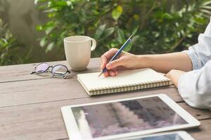 hand kvinna skrivning anteckningsbok på trä tabell med kopp coffe och läsplatta. foto