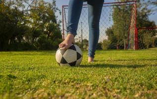 ben av en kvinna i blå byxor, barfota i de mitten av de parkera spelar med en fotboll boll med en fotboll mål med nät och träd i de bakgrund under de morgon- foto