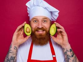 Lycklig kock med skägg och röd förkläde innehar ett avokado foto