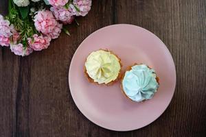 en topp se av två gul och blå grädde capcakes på en rosa tallrik sitta på en trä- tabell. en beige kopp av cappuccino sitter på de tabell. nära en tallrik av kakor lögn skön rosa och vit blommor. foto