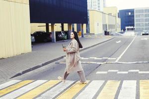 kvinna innehav kaffe och korsning de väg på fotgängare korsning mot urban stad bakgrund foto