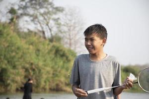 asiatisk Pojkar håll badminton fjäderboll och racket, stående och spelar bredvid de flod Bank i deras lokal- flod under deras helgen Semester, mjuk och selektiv fokus på främre pojke i vit skjorta. foto