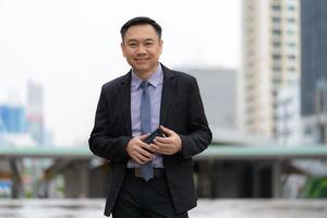 asiatisk affärsman som står och håller mobiltelefonen med kontorskontorsbyggnader i stadsbakgrunden foto