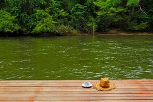 ta en koppla av tid med kopp av kaffe och sugrör hatt på brun trä- golv med suddig flod och grön skog bakgrund på semester dag på kanchanaburi, thailand- skönhet av natur och landmärke begrepp foto