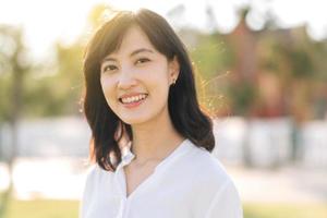 porträtt ung skön asiatisk kvinna med Lycklig leende runt om utomhus- parkera i solig sommar dag foto